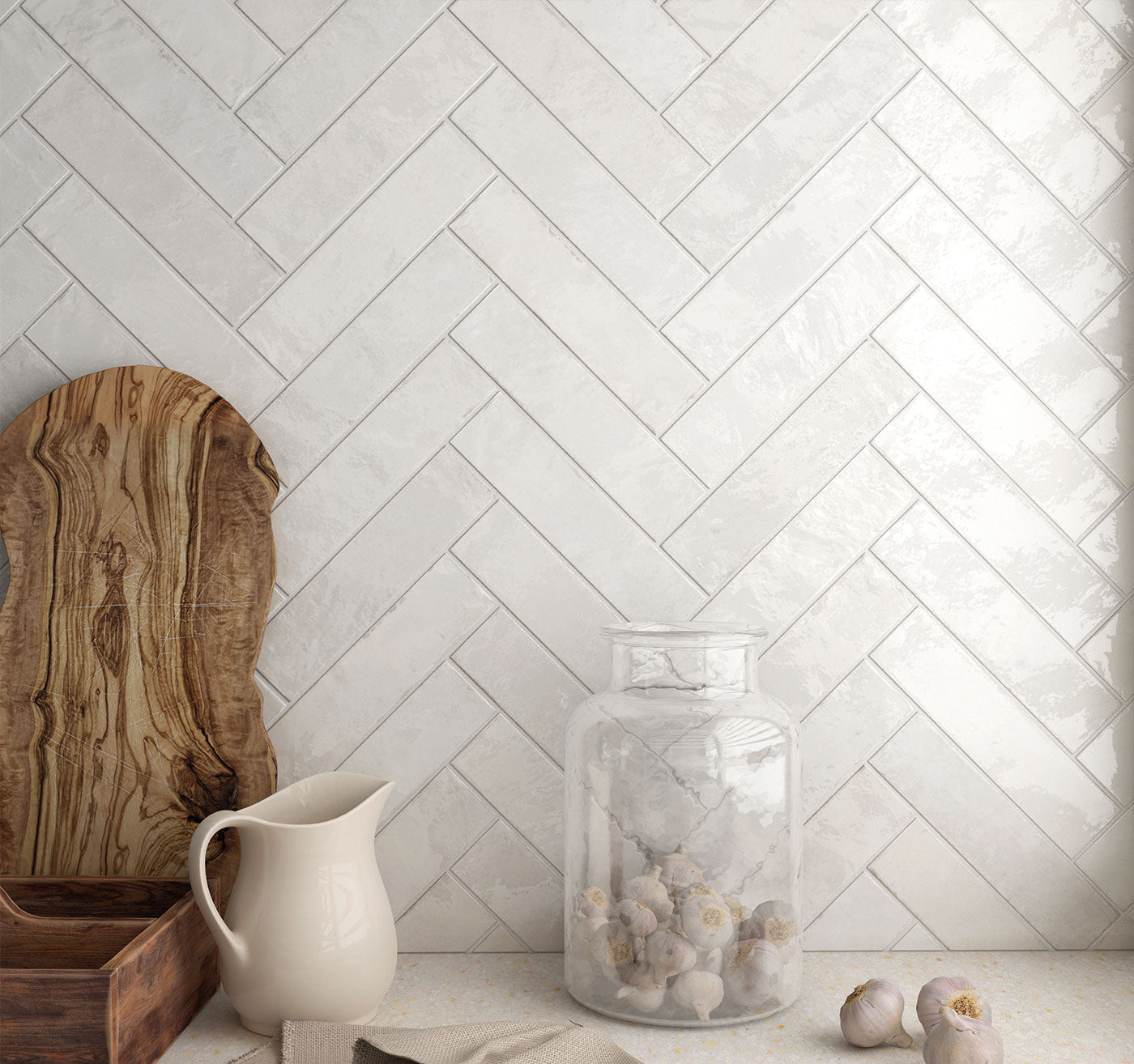 White tiled wall with a herringbone pattern, wooden cutting board, white pitcher, glass jar, and decorative items.